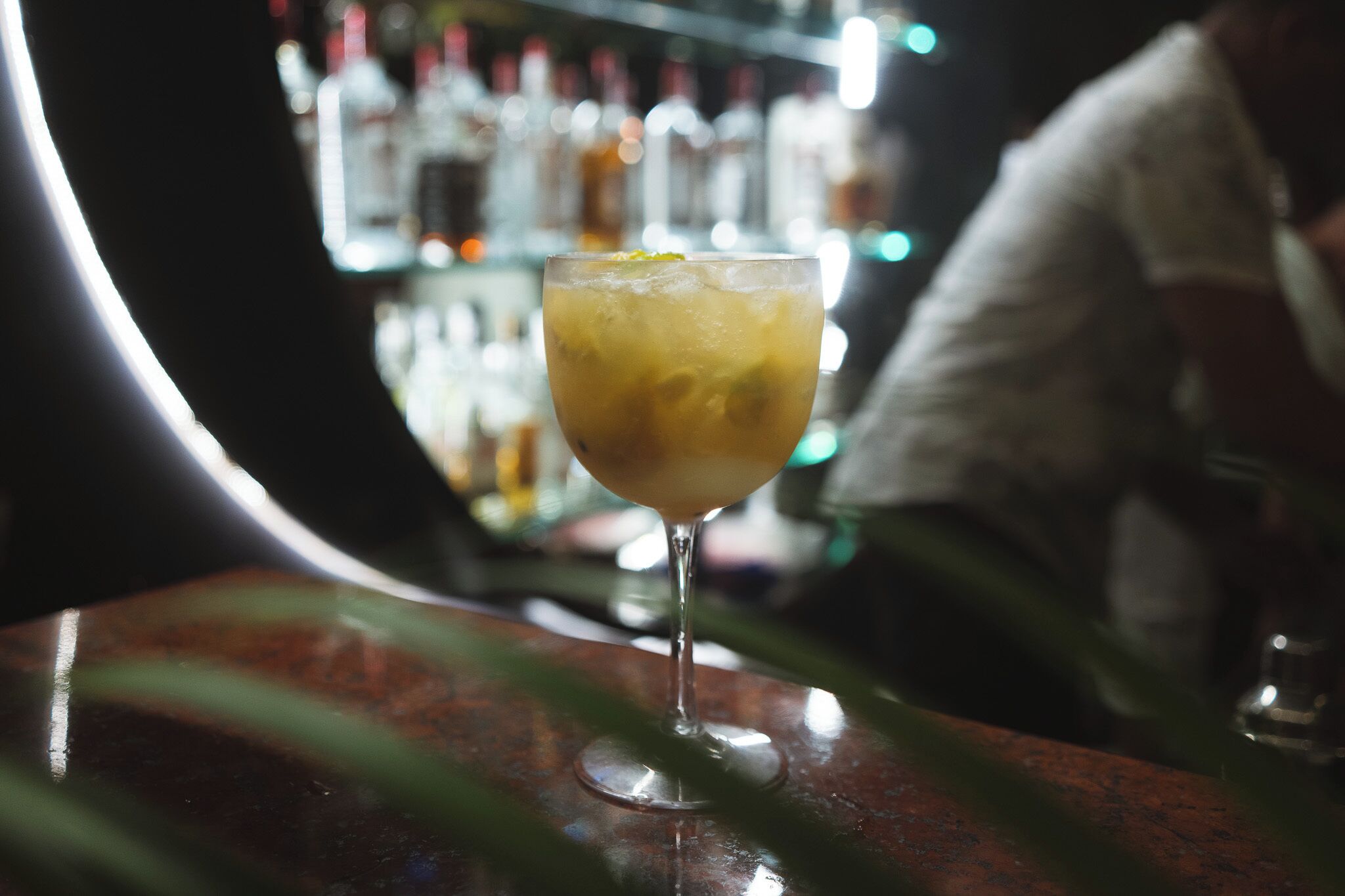 Eine Bar mit einem Cocktailglas auf einer Holztischplatte, im Hintergrund unscharf Regale mit Flaschen und eine teilweise sichtbare Person.