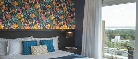 Une chambre d’hôtel avec un grand lit, une table de chevet, une lampe fixée au mur et une vue sur l’extérieur grâce à une grande fenêtre.