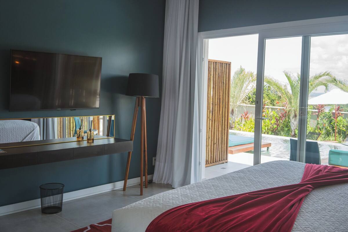 A bedroom with a bed, a TV, a bedside table, and a window with a view of a pool and greenery.