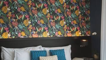 Una cama con ropa de cama blanca y azul, cabecera oscura y un vibrante papel tapiz tropical.