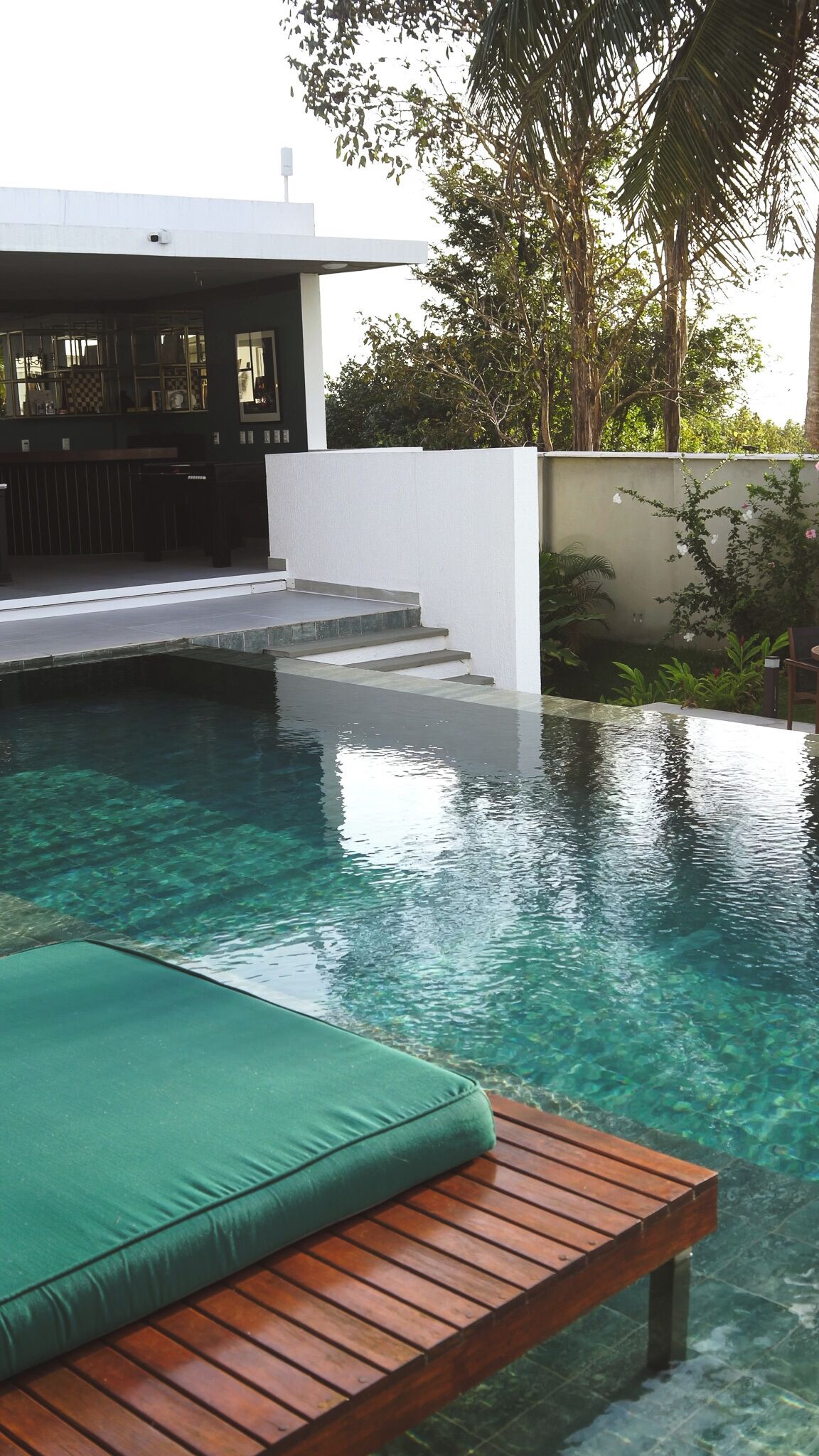 A modern outdoor pool area with clear water, a wooden deck, and a green cushioned lounge chair.