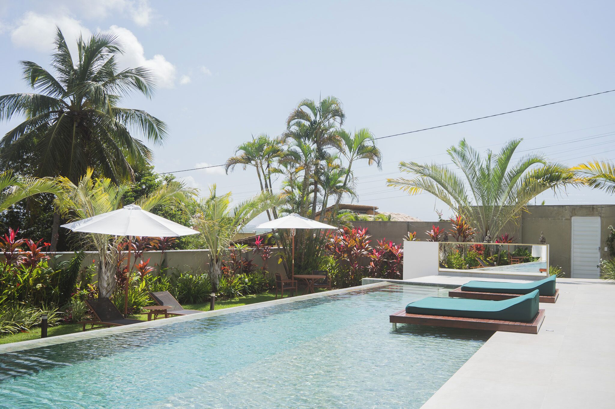 Piscina com espreguiçadeiras e guarda-sóis, cercada por palmeiras e plantas tropicais.