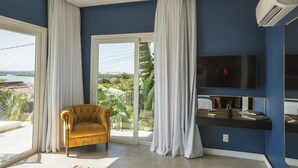 A room with a large window, a yellow armchair, a flat-screen TV, and a sliding glass door leading to a balcony with a view of the beach. - The View Pipa (Tibau do Sul)