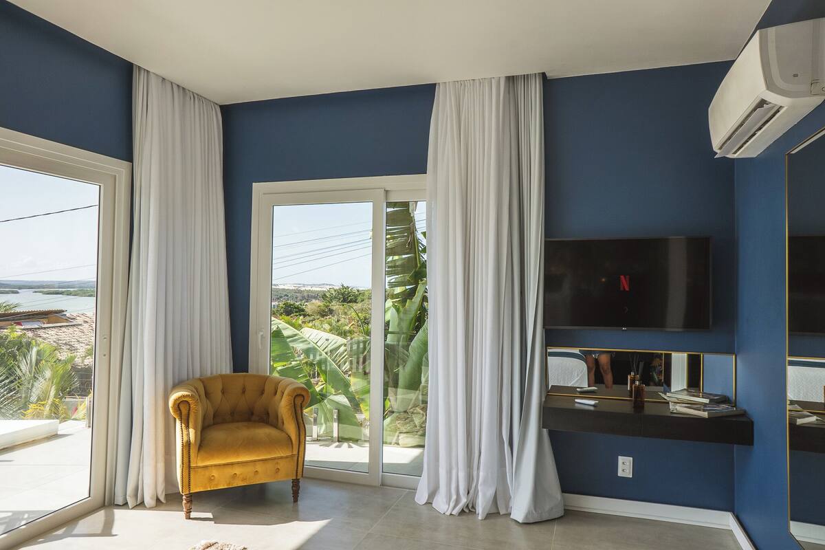 A room with a large window, a yellow armchair, a flat-screen TV, and a sliding glass door leading to a balcony with a view of the beach.