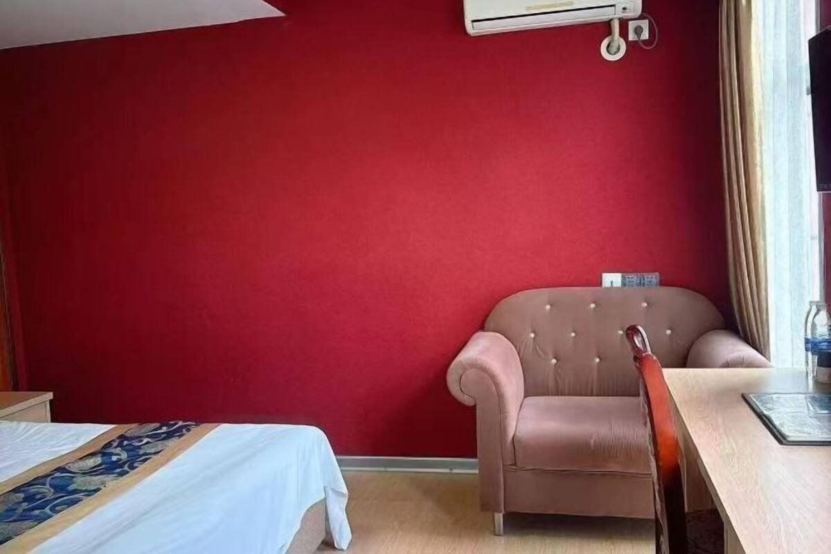 Habitación de hotel con una pared roja, una cama con edredón estampado, un sillón color beige, una mesa de madera y un equipo de aire acondicionado.
