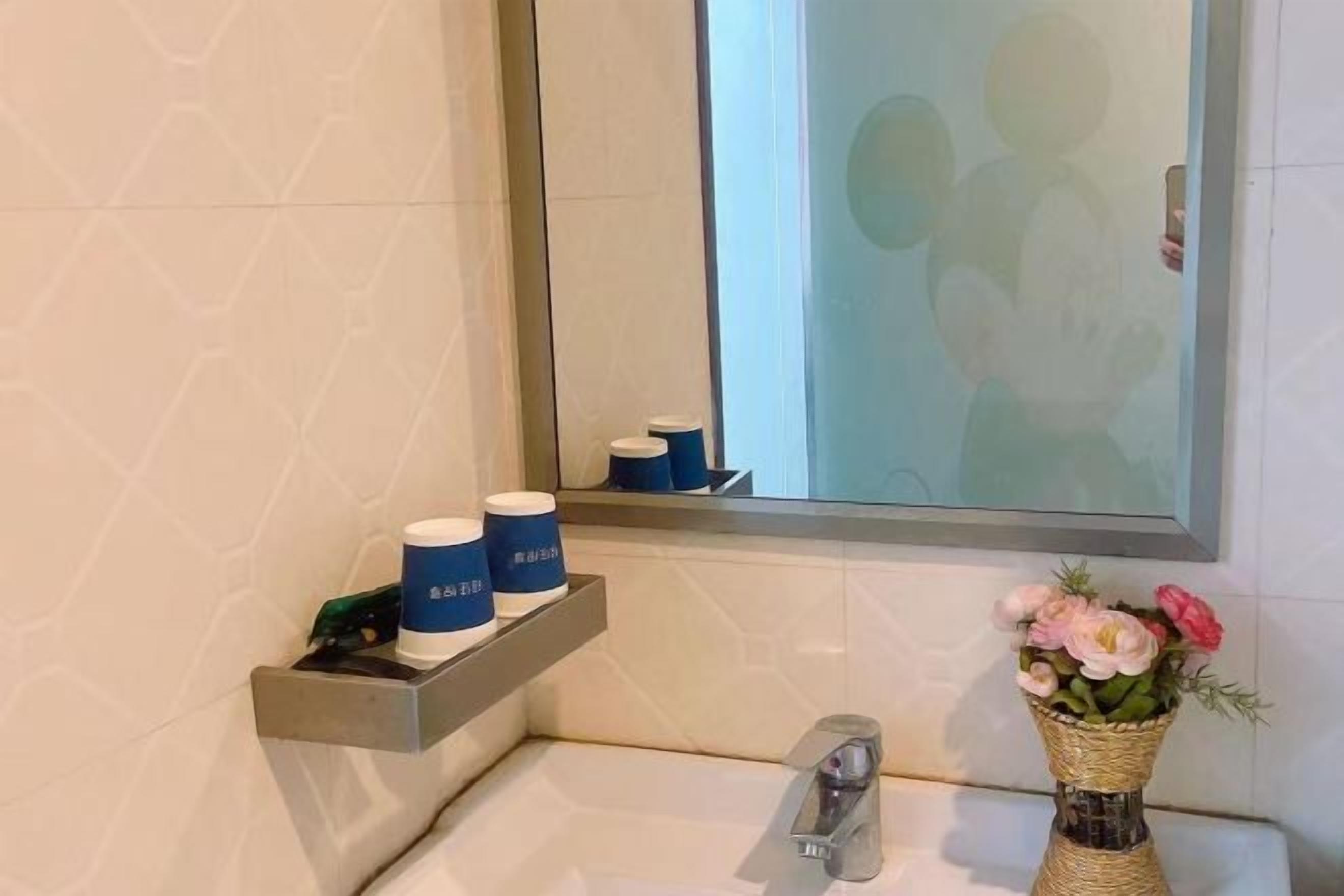 A bathroom with a mirror, a shelf with two bottles, a vase with flowers, and a sink.