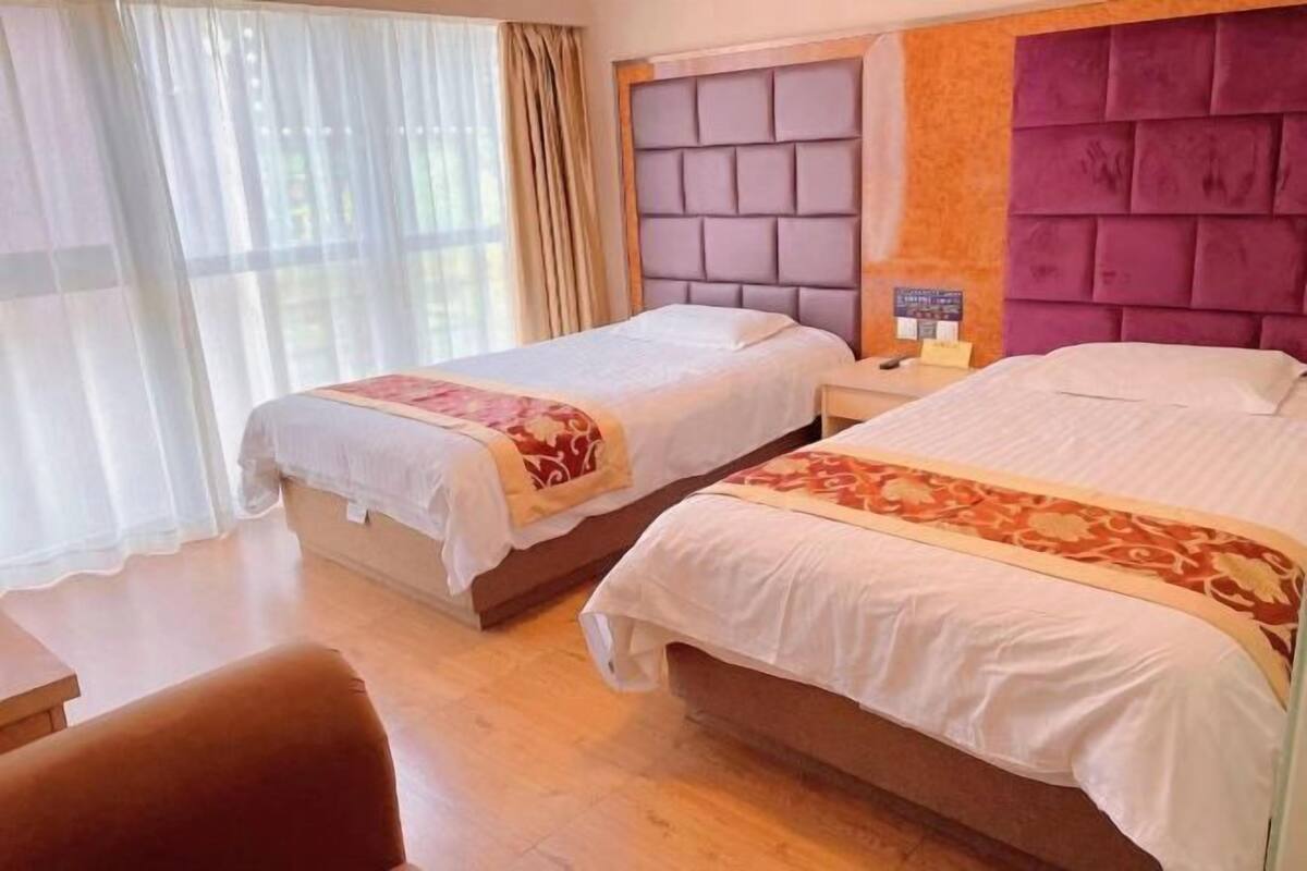 Habitación de hotel con dos camas, piso de madera, ventana con cortinas translúcidas y mesita de noche con lámpara.