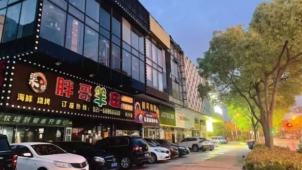 A multi-story building with large windows and illuminated signs, parked cars along the street, and trees in the foreground. - Light Stay Hotel - Xiaozhu (Shanghai Chuansha Branch) (Shanghai)