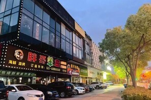 A multi-story building with large windows and illuminated signs, parked cars along the street, and trees in the foreground.
