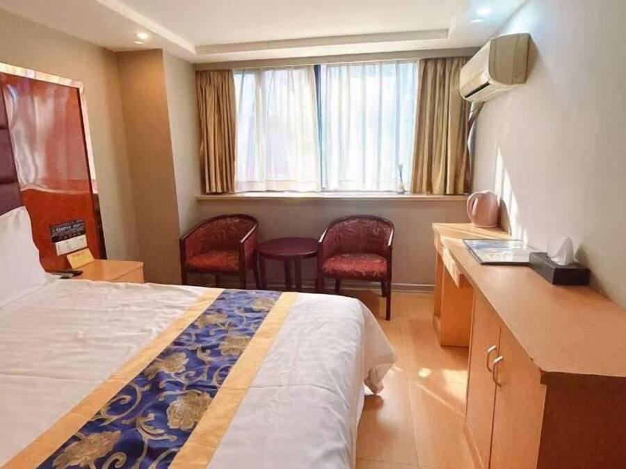 Quarto de hotel com uma cama grande, uma escrivaninha, duas cadeiras, uma janela com cortinas e um ar condicionado fixado na parede.