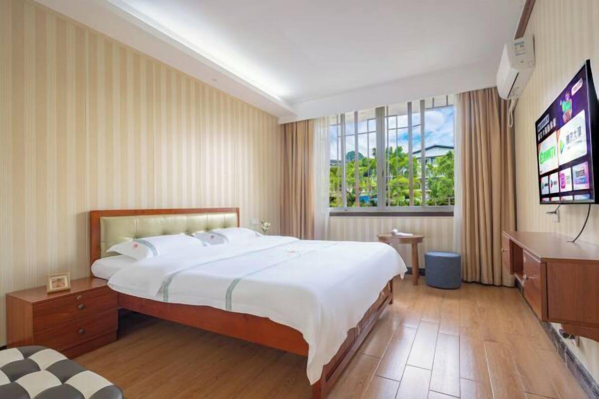 A hotel room with a large bed, a TV, a desk, and a window with a view of greenery.