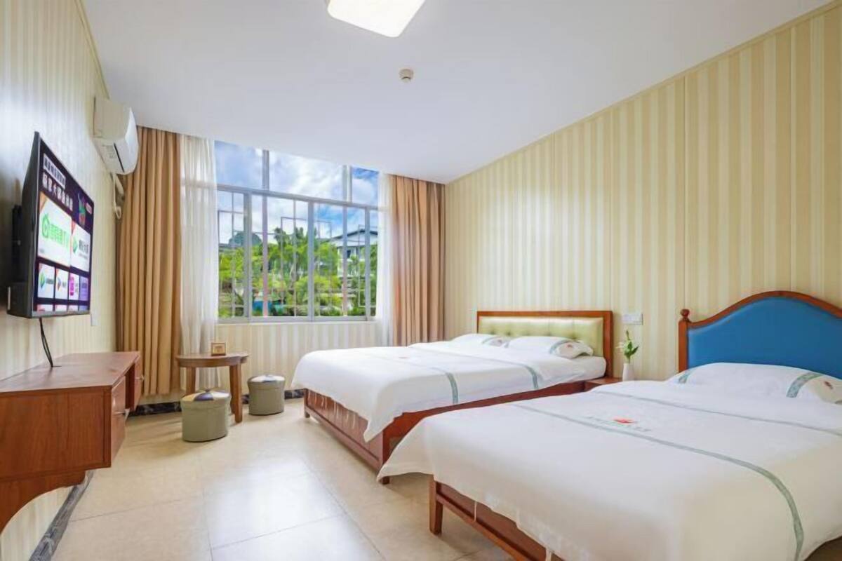 A hotel room with two beds, a TV, a small table, and a window with a view of greenery.