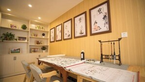 Ein Zimmer mit einem langen Tisch, der Kalligraphie und Gemälde zeigt, Holzregalen mit gerahmten Bildern und einem traditionellen Tuschegemälde an der Wand.