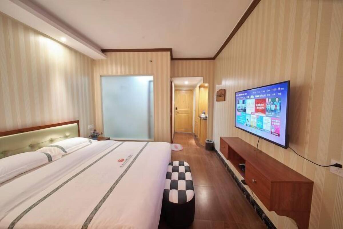 A hotel room with a bed, a television on the wall, a wooden bench, and a door leading to another room.