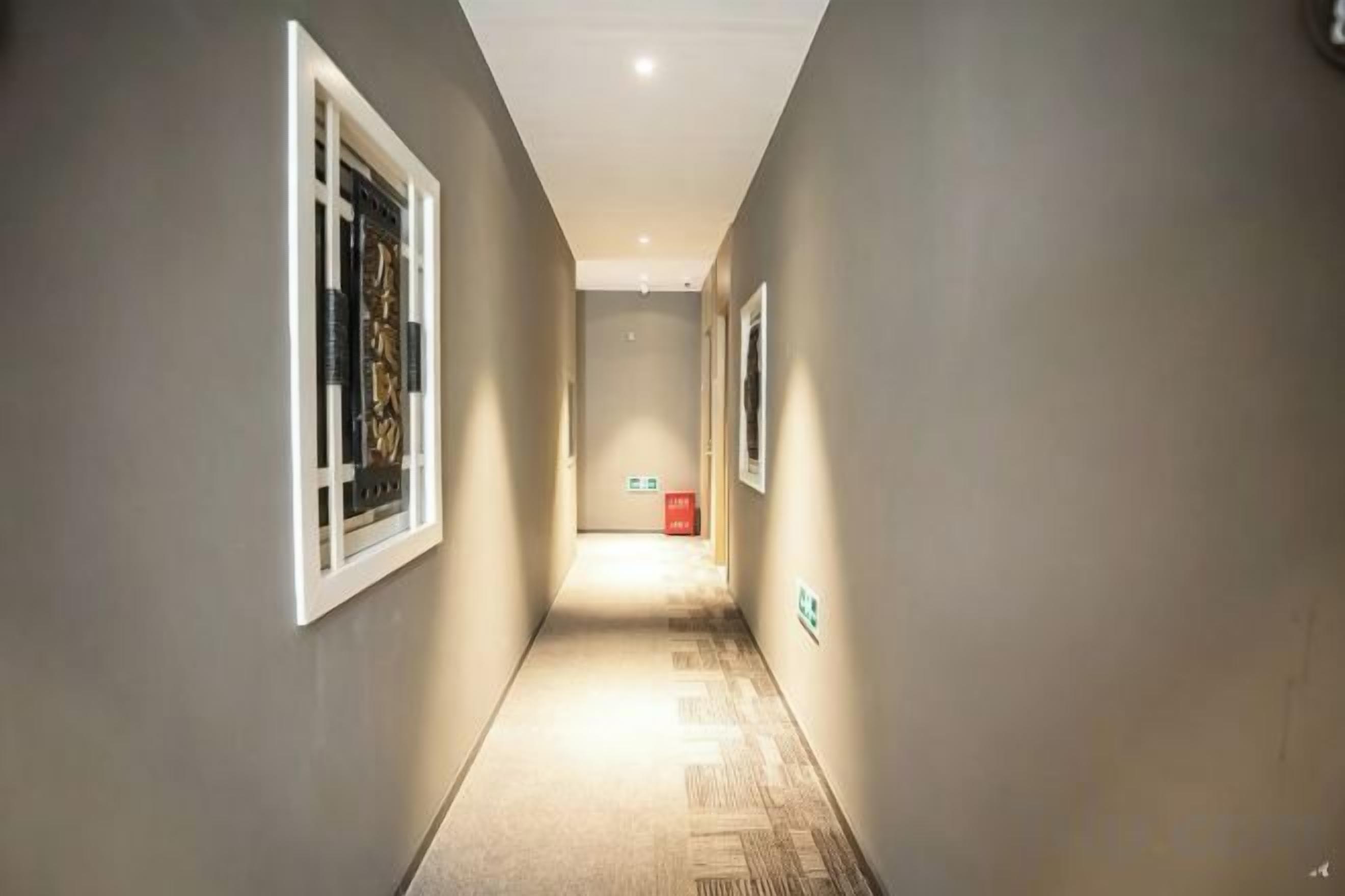 A narrow hallway with a carpeted floor, white-framed windows, and a red emergency exit sign.