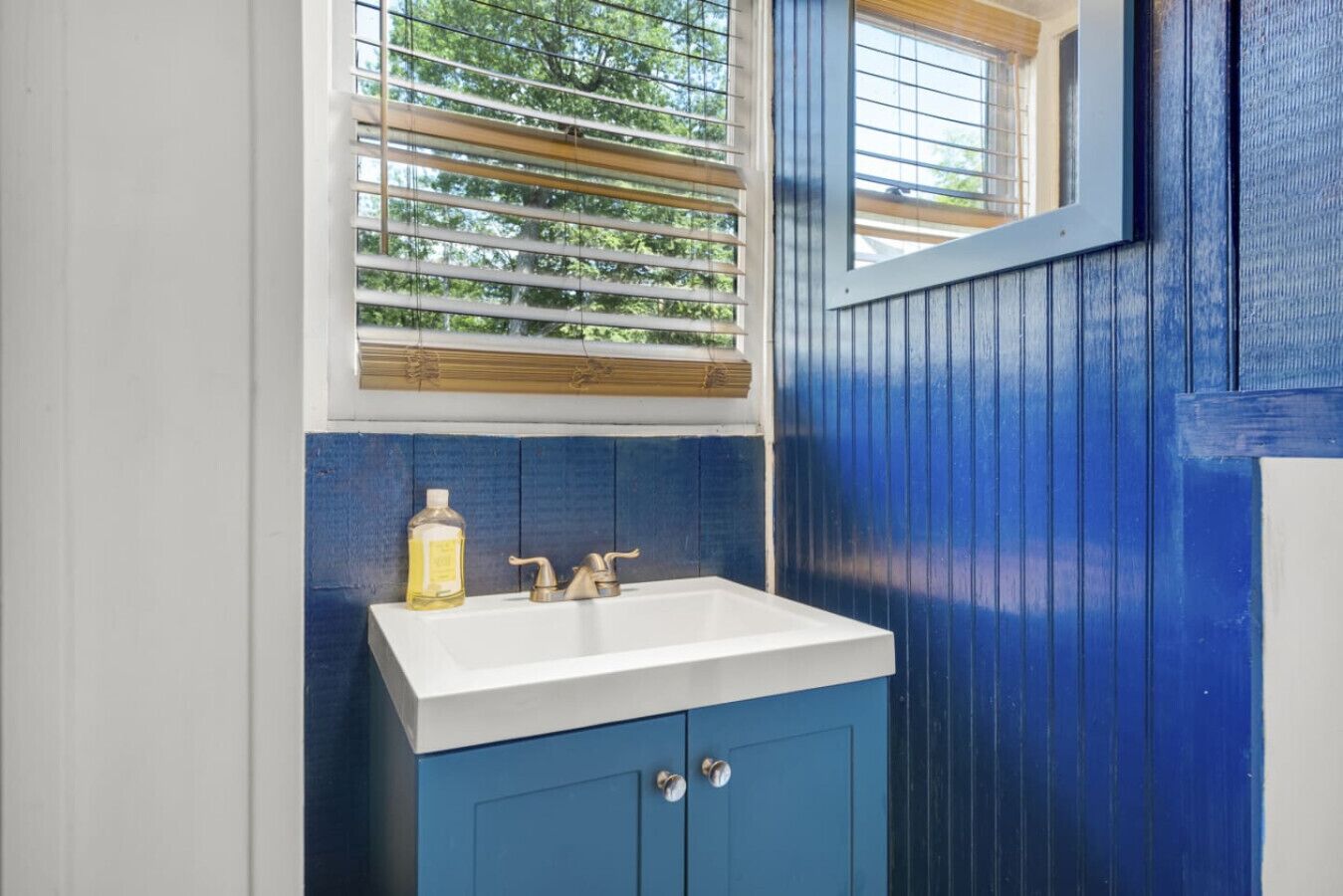 Ein Badezimmer mit einem weißen Waschbecken, blauen Schränken und einem Fenster mit Jalousien.