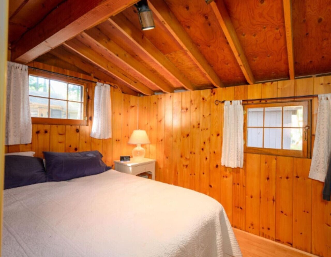 Ein Schlafzimmer mit Holzvertäfelung, einem Bett, einem Nachttisch, einer Lampe und zwei mit Vorhängen versehenen Fenstern.