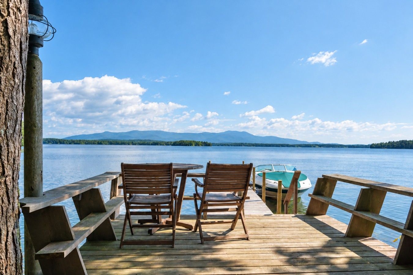 A wooden deck with two wooden chairs and a small round table, overlooking a calm lake with a boat docked nearby.