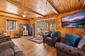A cozy cabin interior with a fireplace, leather sofas, and a large window overlooking a scenic landscape.