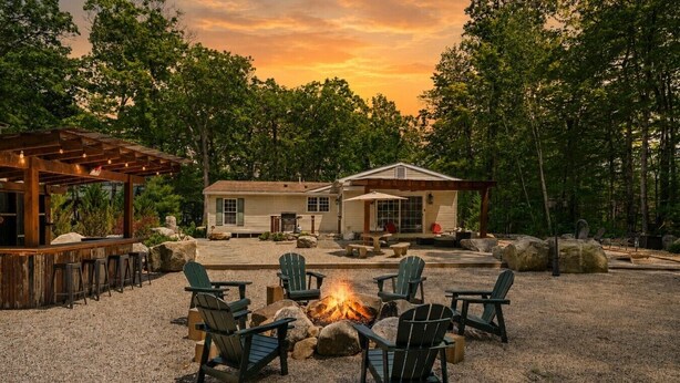 A rustic outdoor seating area with Adirondack chairs around a fire pit, a wooden pergola, and a house with a porch. - Peaceful Escape | Dock Option + Fire pit | Winni (Moultonborough)