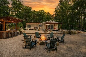 A rustic outdoor seating area with Adirondack chairs around a fire pit, a wooden pergola, and a house with a porch. - Peaceful Escape | Dock Option + Fire pit | Winni (Moultonborough)