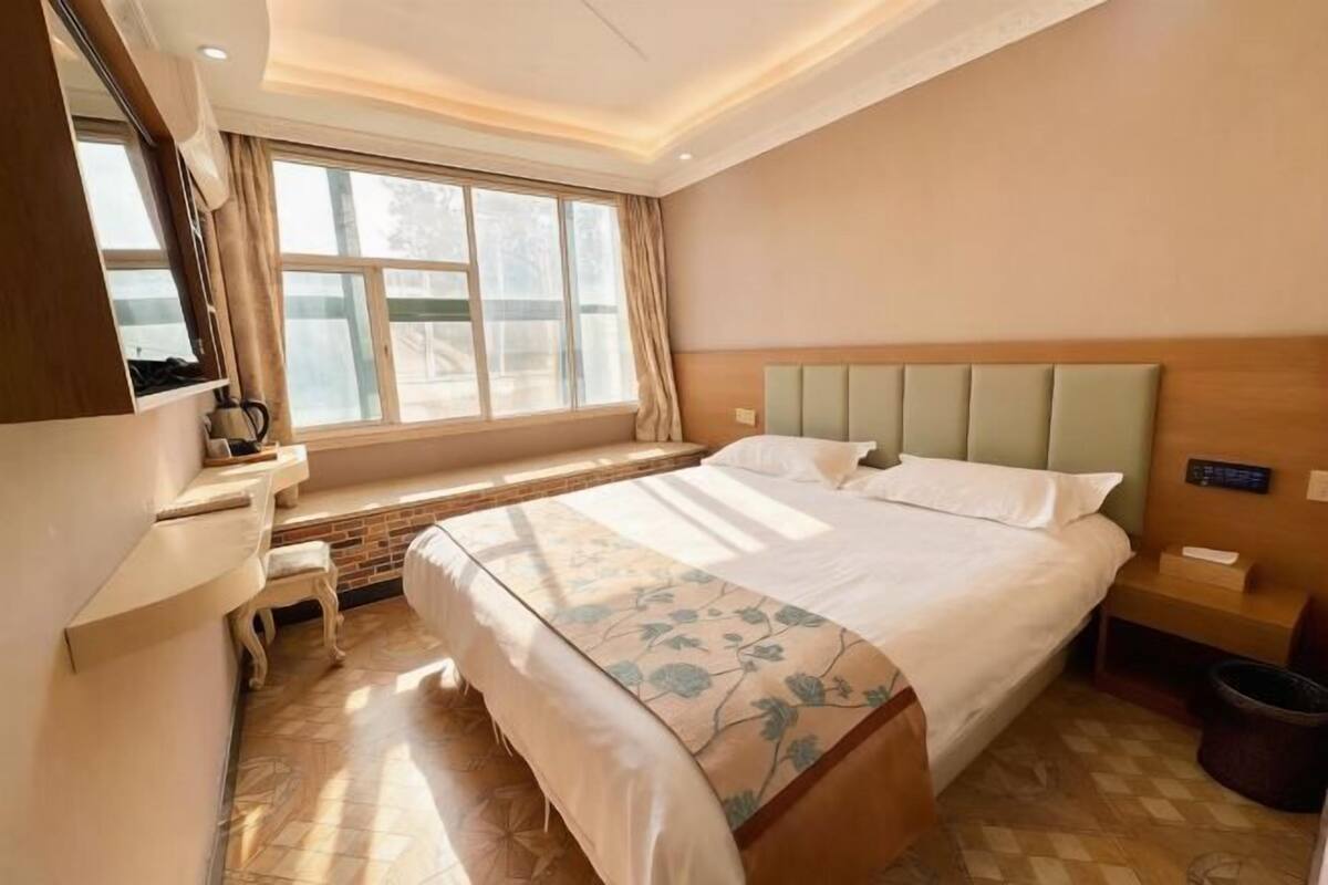 A hotel room with a bed, a desk, a chair, a window, and a trash bin.