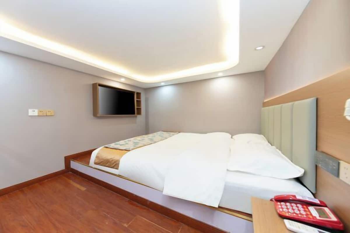 A hotel room with a bed, a bedside table with a phone, a TV mounted on the wall, and a small shelf.