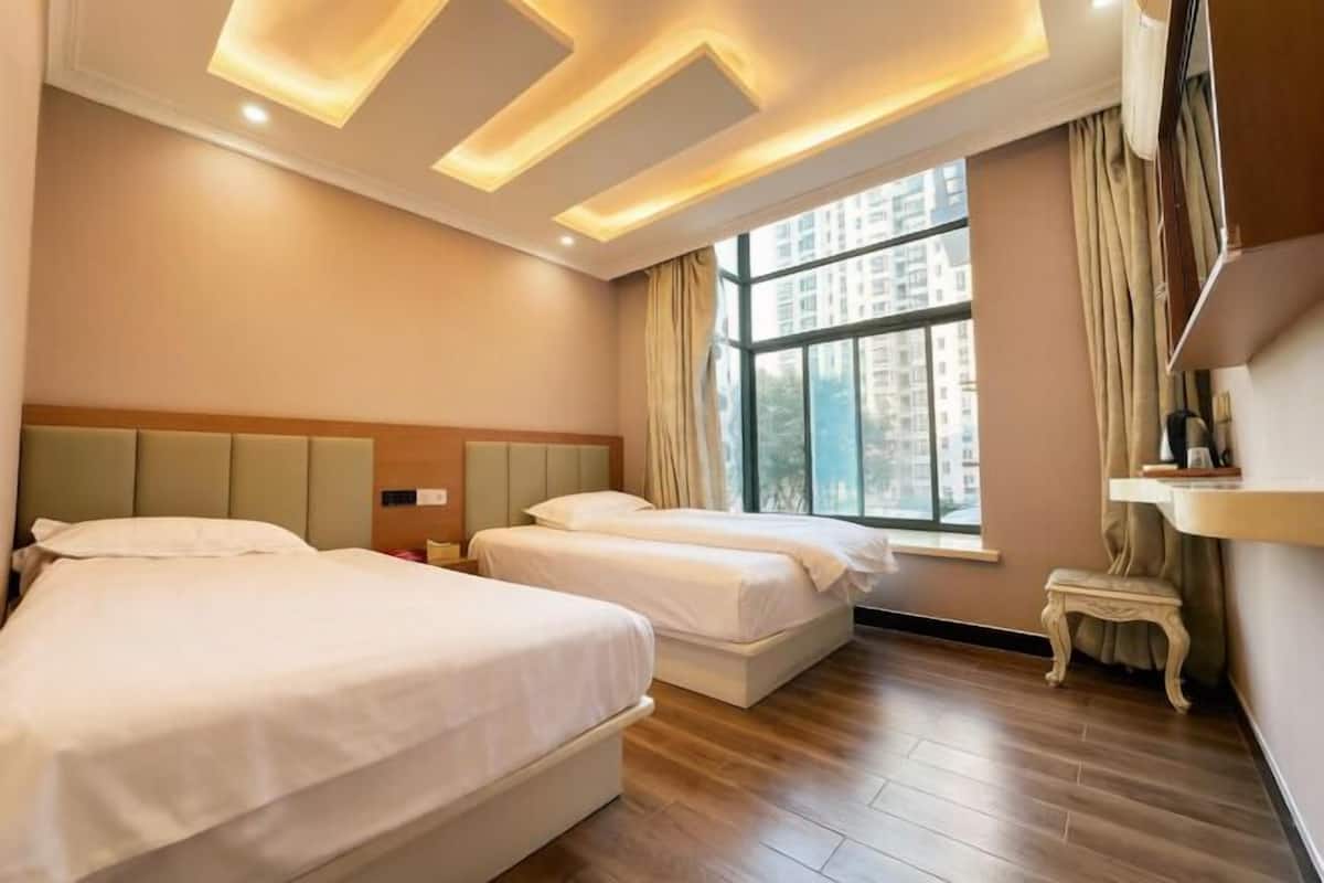 A hotel room with two beds, a small table, a window with a view of buildings, and a ceiling with recessed lighting.