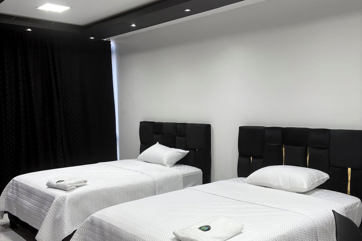 A hotel room with two beds, each with a white quilted bedspread and a pillow. The room has a modern design with a black headboard and a white ceiling.