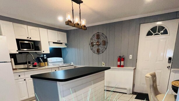 A modern kitchen with white cabinets, a black countertop, and a central island. There is a clock on the wall and a fire extinguisher near the door. - The Suite at North Shore Long Island, Prime Location, Unique and sophisticated (Smithtown)
