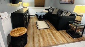 A living room with a black sofa, a wooden coffee table, a black side table, a lamp, a mounted TV, and a window with blinds. - The Suite at North Shore Long Island, Prime Location, Unique and sophisticated (Smithtown)
