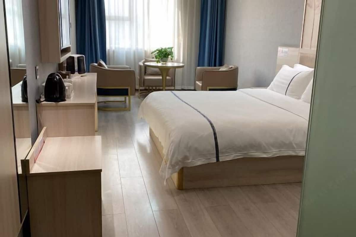 A hotel room with a bed, a desk, a chair, a small table, and a potted plant.