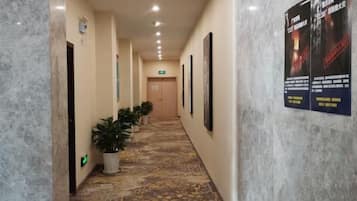 A hotel corridor with a patterned floor, potted plants, framed pictures on the walls, and a poster advertising a hotel event.