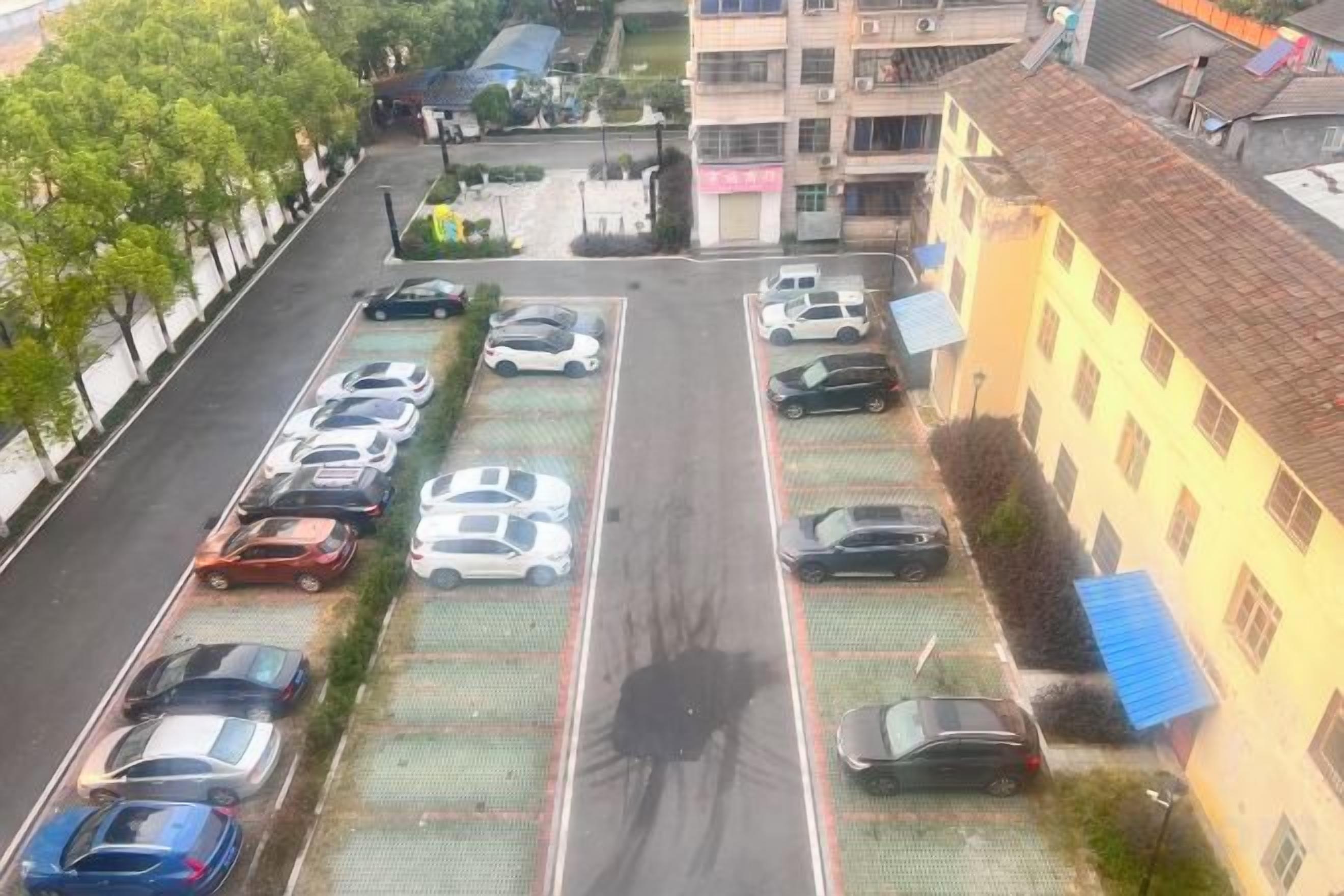 A parking lot with multiple rows of parked cars, a building with a tiled roof, and a tree-lined street.