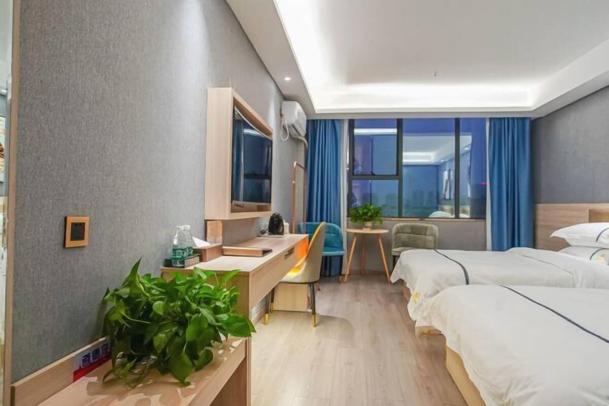 A hotel room with a desk, two beds, a chair, a small table, and a potted plant.