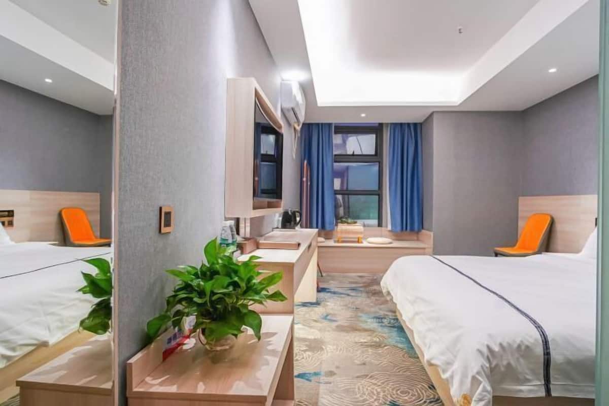 A hotel room with two beds, a desk, a chair, a plant, and a window with curtains.