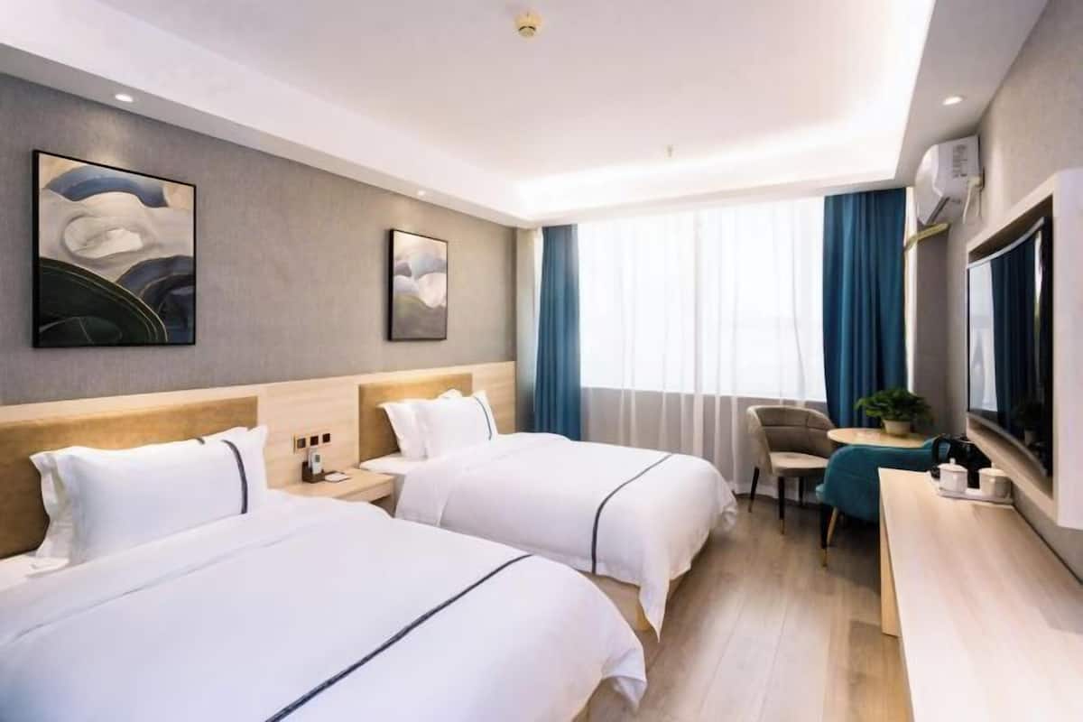 A hotel room with two beds, a desk, a chair, a small table with a plant, and artwork on the walls.