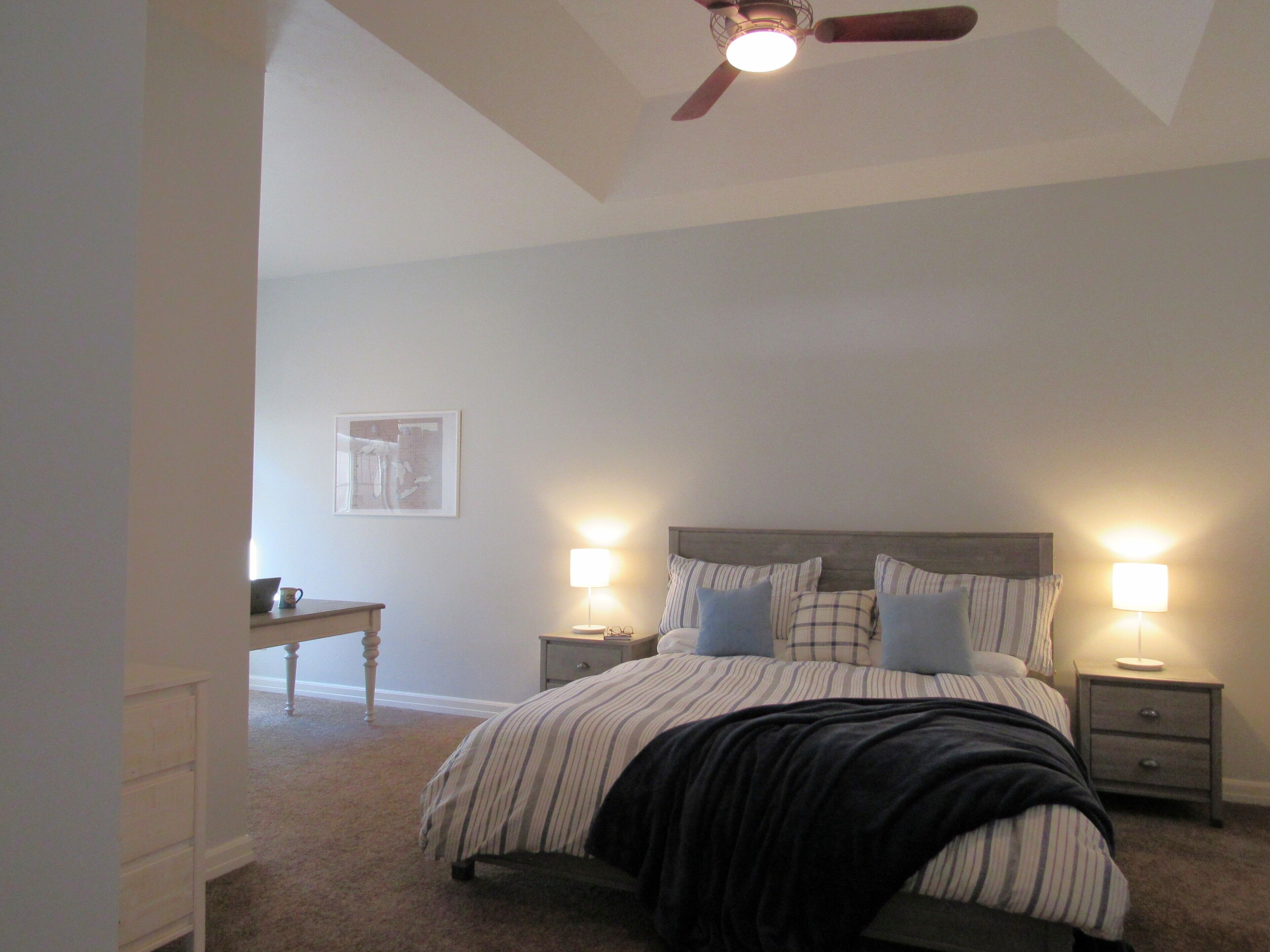 A bedroom with a bed, two bedside tables with lamps, a nightstand, a desk, and a ceiling fan.