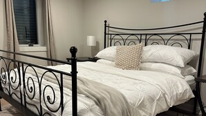 Una cama bien hecha con ropa blanca, un cojín decorativo estampado y un cabecero de metal negro. Hay una ventana con cortinas, una mesita de noche con lámpara y una alfombra estampada en el suelo.