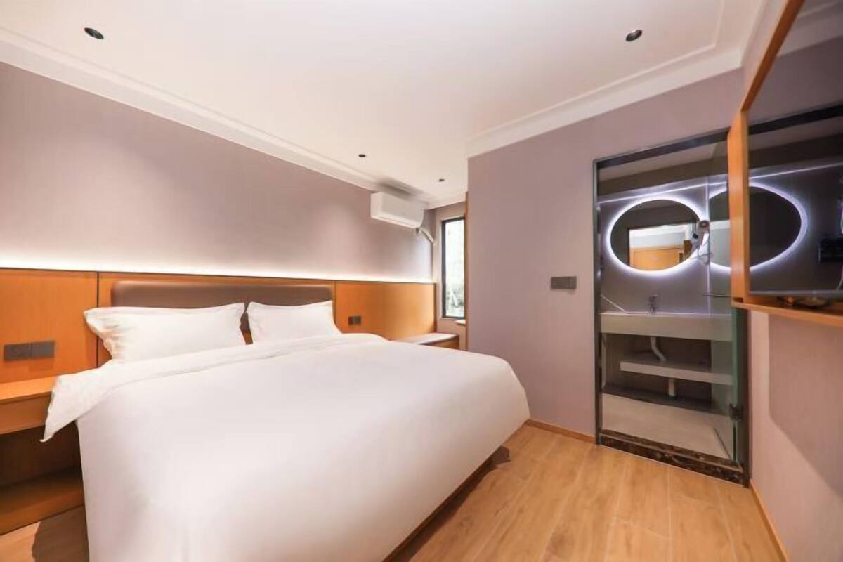 A modern hotel room with a large bed, a wooden headboard, a built-in wardrobe with a mirror, and a small bedside table.