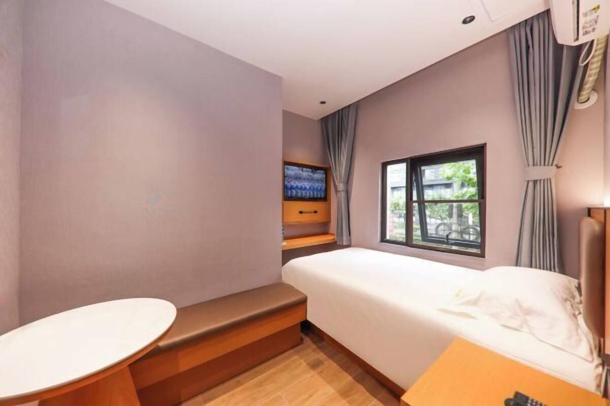 A hotel room with a bed, a small table, a window with curtains, and a view of greenery outside.