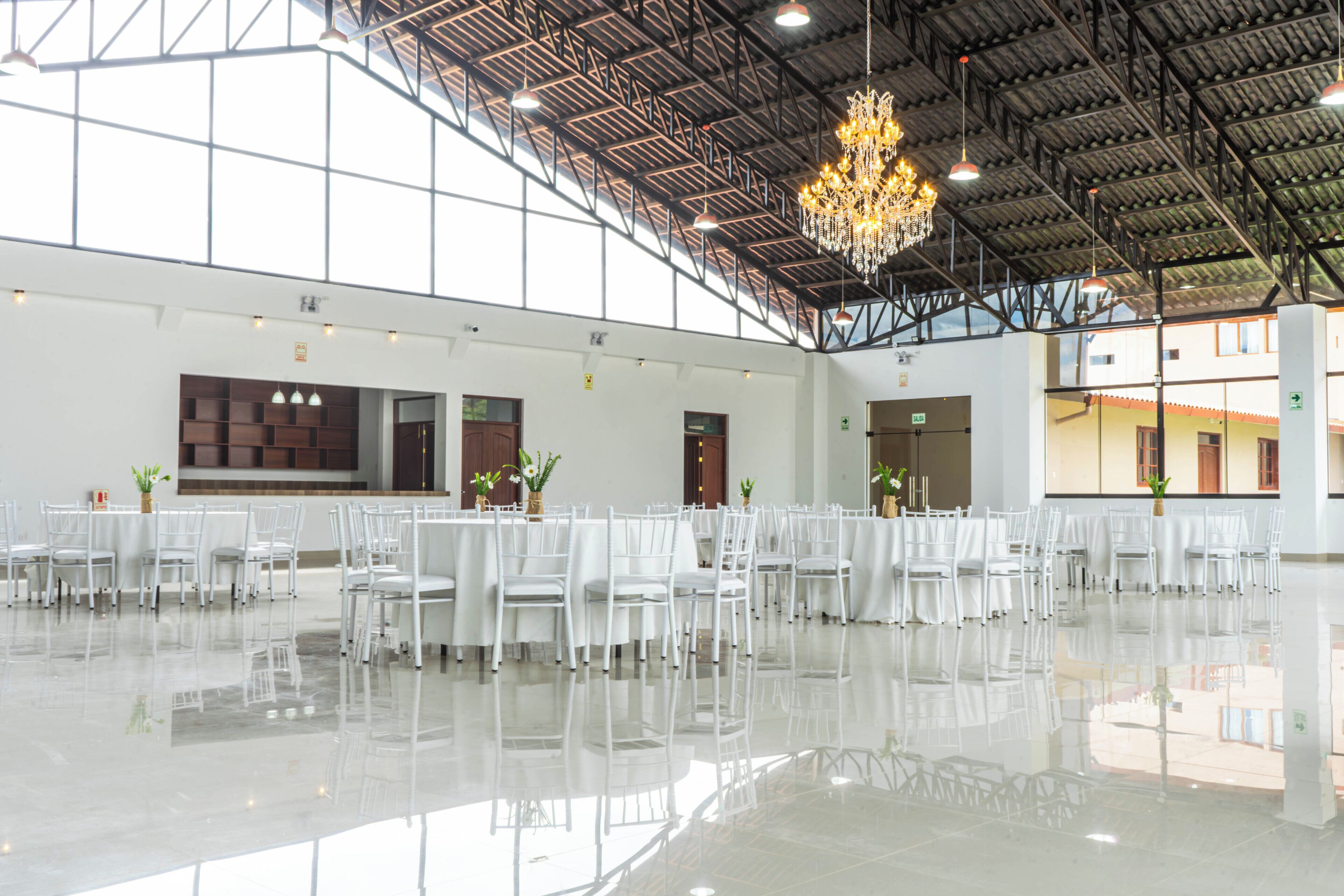 Ein geräumiger Saal mit weißen Tischen und Stühlen, einem großen Chandelier und einer hohen Decke mit Metallbalken.