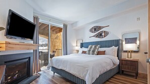 Une chambre avec une cheminée, un lit avec une literie à rayures, un téléviseur fixé au-dessus de la cheminée et un balcon offrant une vue sur un paysage enneigé.
