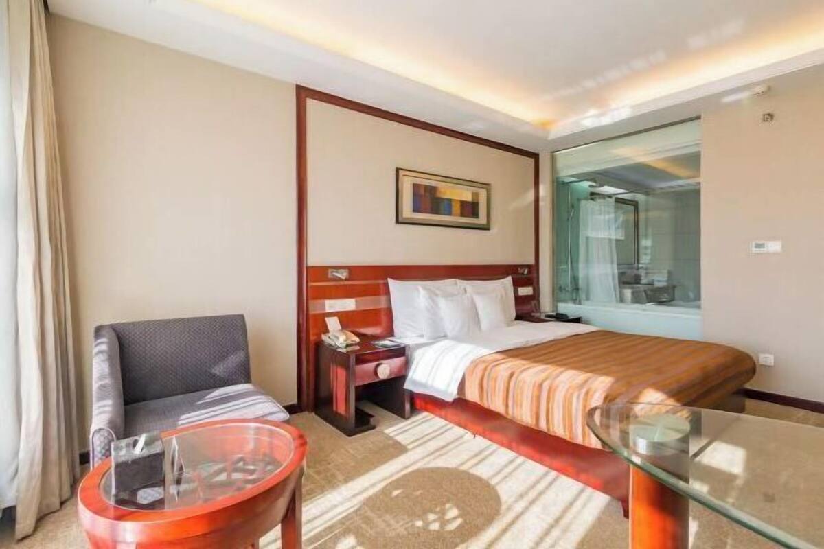 A hotel room with a bed, a chair, a glass table, and a nightstand.
