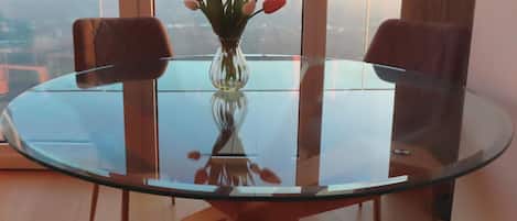 A round glass table with a vase of flowers in the center, surrounded by two chairs, in a room with large windows and a view of the city.