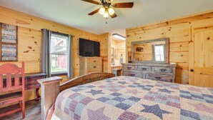 A cabin-style bedroom with a bed, wooden walls, a ceiling fan, a dresser, a television, and a window with a view of a house. - Fire Pit + Game Room: Lakefront Escape in Trenton! (Trenton)
