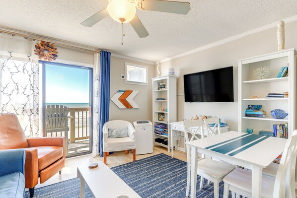 A living room with a TV, a table, chairs, a bookshelf, and a view of the ocean through a sliding door. - Oceanfront Gem w/ Views in North Topsail Beach (North Topsail Beach)
