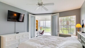 A bedroom with a large bed, a TV mounted on the wall, a ceiling fan, and a view of the outdoors through a window. - Walk to Stuart Beach: Resort Condo w/ Water View! (Stuart)