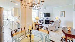 A modern kitchen with white cabinets, a dining area with a glass table, and a living room with a TV and seating. - Walk to Stuart Beach: Resort Condo w/ Water View! (Stuart)