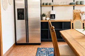 Una cocina con un refrigerador de acero inoxidable, una cafetera y una mesa de comedor de madera con una silla.